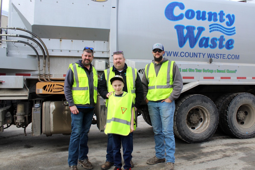 Three men and boy wearing safety vest in front of garbage truck