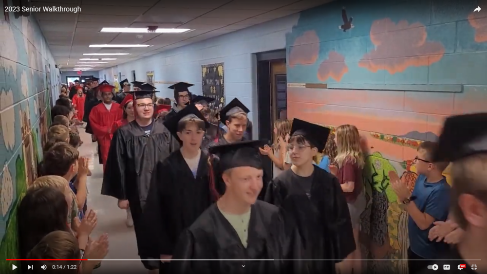 Seniors in caps and gowns walk through hallway lined with elementary school students