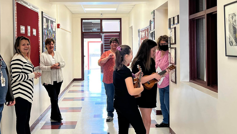 two students playing ukuleles for staff members in hall