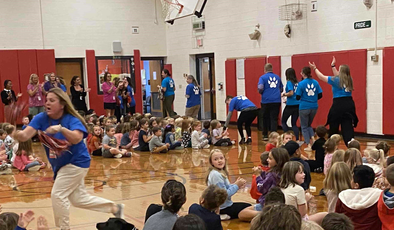Teachers in blue shirts in front of audience of children sitting on gym floor