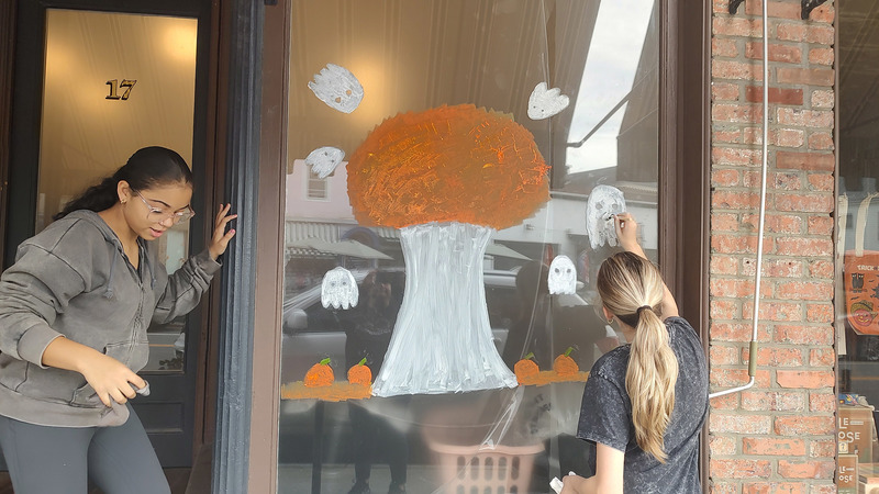 two girls painting tree surrounded by ghosts on shop window.